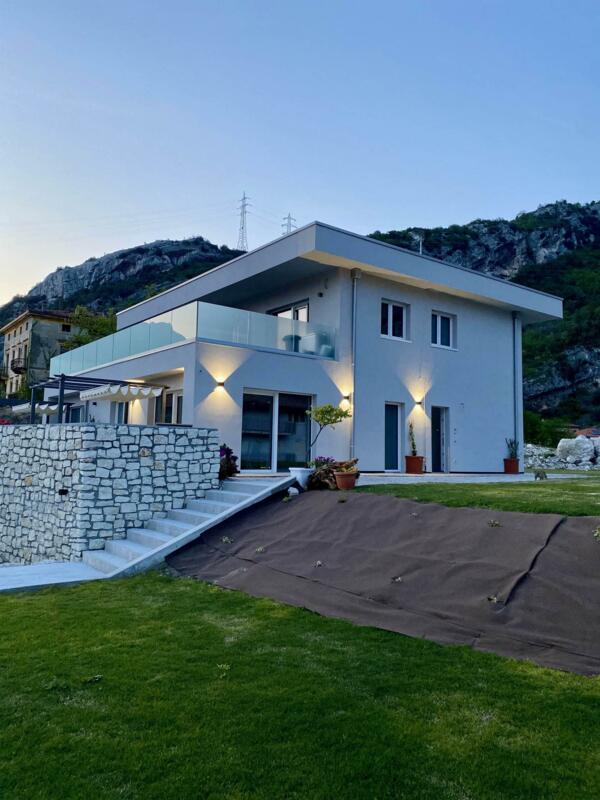 Eine moderne weiße Villa mit Steindetails, umgeben von einem großen Garten. Im Hintergrund sind Hügel und ein klarer Himmel bei Sonnenuntergang zu sehen.