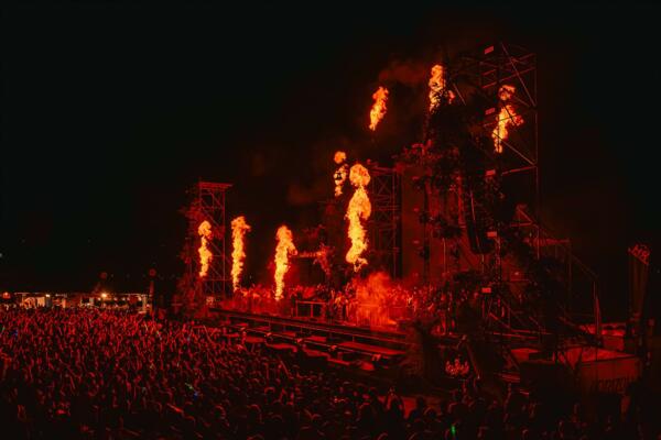 Un grande concerto con una folla entusiasta. Si vedono spettacolari effetti di fiamme e luci sullo sfondo scuro.