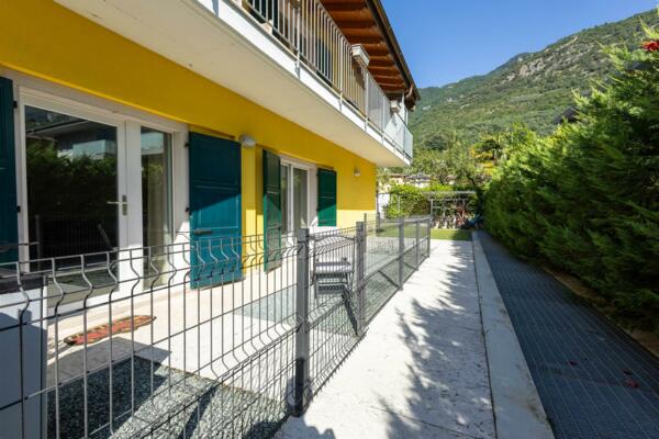 Eine moderne Villa mit gelben Wänden und grünen Fenstern. Der Garten ist gepflegt, von einem Zaun umgeben, mit Bergen im Hintergrund.