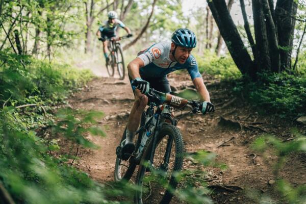 Due ciclisti sono in gare su un sentiero di terra circondato da alberi e vegetazione. Il ciclista in primo piano sta affrontando una curva con determinazione.