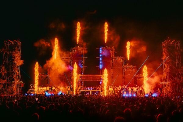 Ein großes Konzert mit einer Bühne, die von Flammen und bunten Lichtern beleuchtet wird. Die begeisterte Menge applaudiert und hält die Telefone hoch, um das Ereignis aufzuzeichnen.