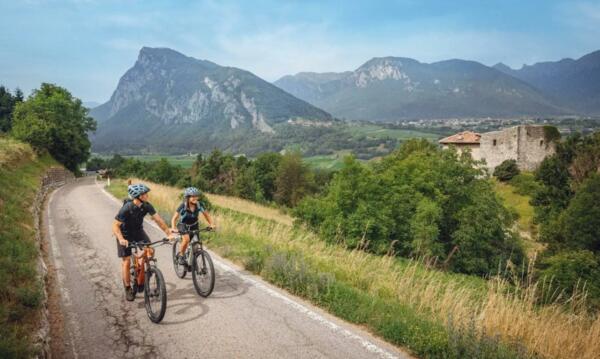 Two cyclists ride along a road surrounded by green hills and majestic mountains. It is a beautiful day outdoors.
