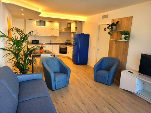 Ein gemütliches modernes Wohnzimmer mit einem blauen Sofa und zwei Sesseln. Die Küche ist ausgestattet und es gibt eine dekorative Pflanze, die einen Hauch von Frische hinzufügt.