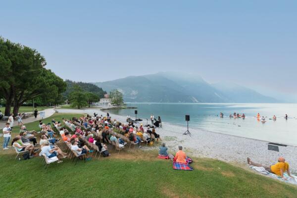 Una folla di persone sedute su sedie assiste a un evento vicino a un lago. Sullo sfondo, si possono vedere le montagne e alcune persone che nuotano.