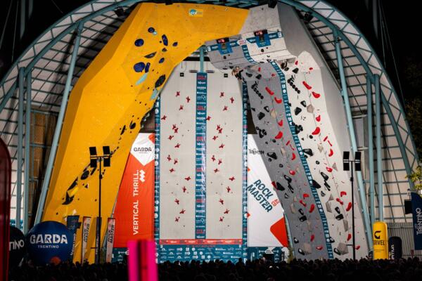 Una struttura di arrampicata con pareti gialle e grigie, allestita per una competizione. Una folla di spettatori si raduna sotto l'evento.