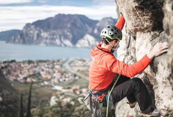 A climber in a red t-shirt climbs a rocky wall. In the background, there is a lake and a town nestled in nature.