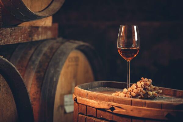 Un bicchiere di vino rosato è appoggiato su una botte di legno, circondato da grappoli di uva essiccata. Dietro, si possono vedere altre botti in un ambiente meravigliosamente rustico.