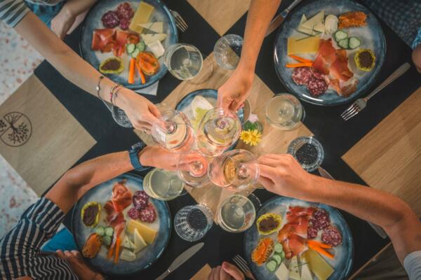 A table laid out with plates of cold cuts, cheeses, and vegetables. A group of people toasts with wine glasses.