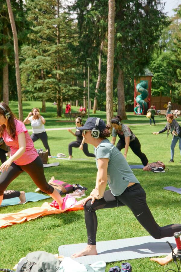 A group of people is participating in an outdoor yoga class, wearing headphones. The scenery is green and surrounded by trees.