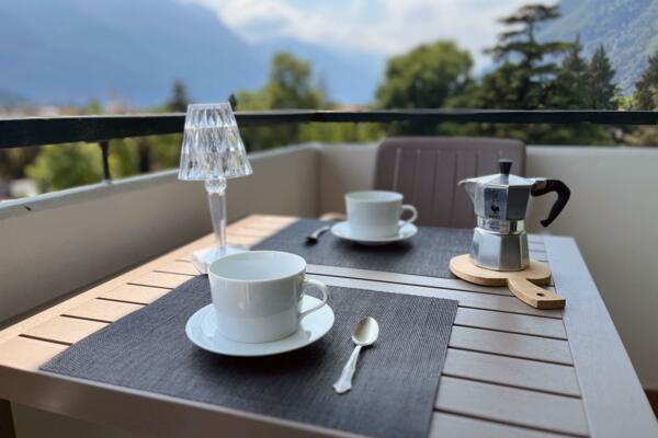 Ein Tisch auf der Terrasse mit zwei Tassen Kaffee und einer Moka. Im Hintergrund sind Bäume und Berge zu sehen.