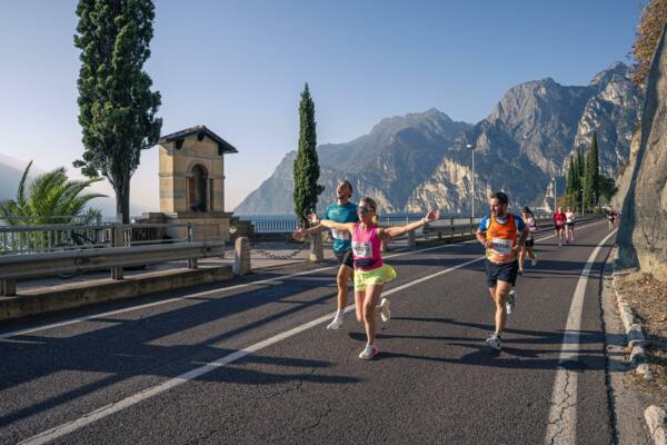 Un gruppo di corridori si sta dedicando a una gara lungo una strada panoramica. Sullo sfondo si vedono montagne e un lago, creando un'atmosfera vivace e naturale.