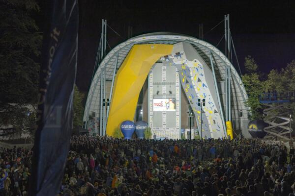 A large climbing competition takes place at night, with a large crowd of spectators. On stage, there are colorful and illuminated climbing walls.