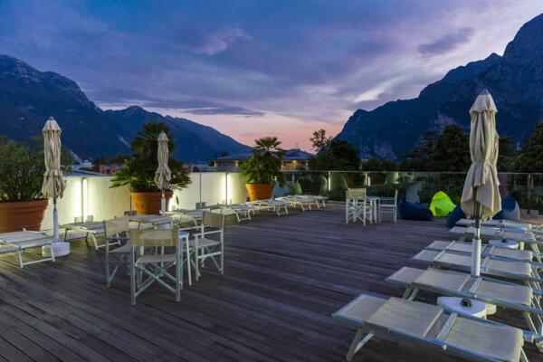 A panoramic terrace with sun loungers and umbrellas, surrounded by plants. In the background, mountains can be seen with a sunset sky.