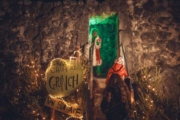 Ein für die Feiertage dekoriertes Haus mit einem Schild "Haus des Grinch". Eine Figur, die sich als Grinch verkleidet hat, heißt die Besucher vor einer beleuchteten Tür willkommen.