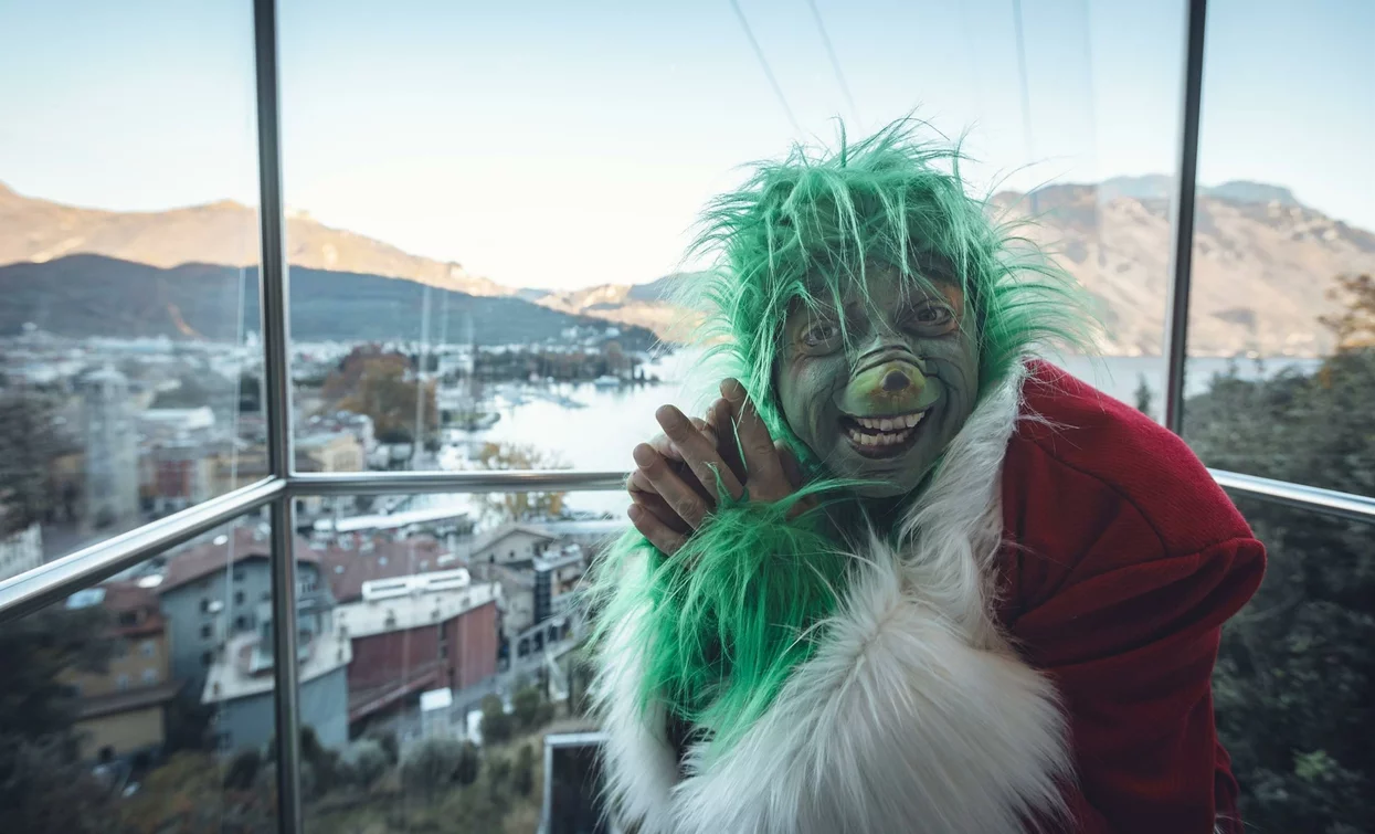Una persona vestita da Grinch, con un viso verde e una giacca rossa, sorride davanti a una vista panoramica di una città e montagne. Il cielo è sereno e chiaro.