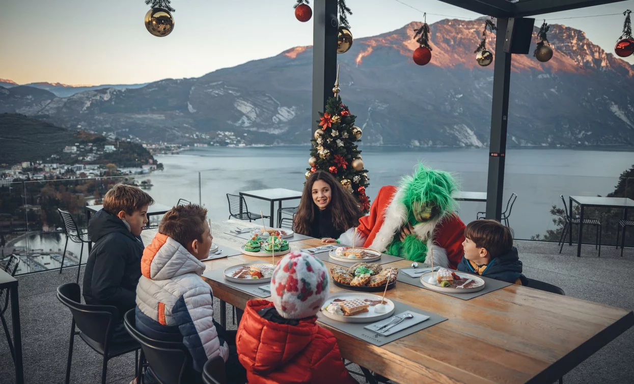 Eine Gruppe von Kindern und eine Figur, die als Grinch verkleidet ist, sitzen an einem Tisch im Freien mit Blick auf einen See und Berge. Sie sind von Weihnachtsdekorationen und Tellern mit Süßigkeiten umgeben.