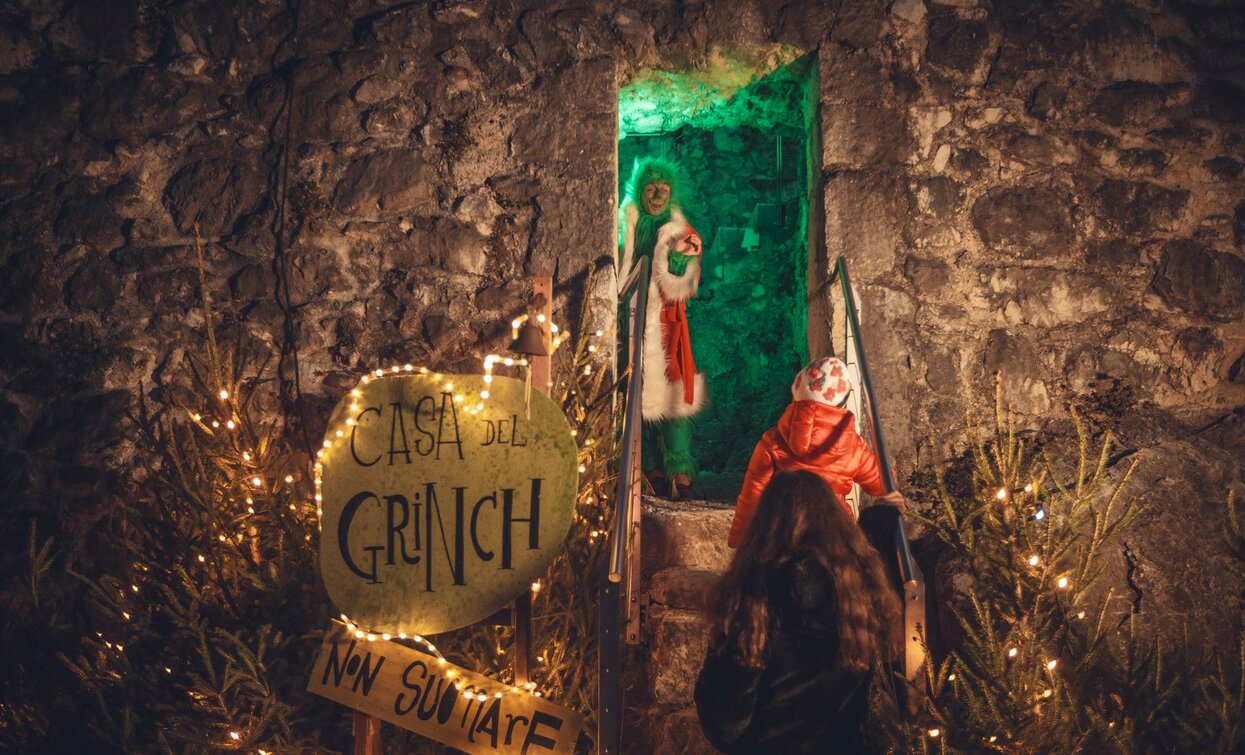 Una casa decorata a tema "Grinch" con luci natalizie. Un personaggio vestito da Grinch si trova sulla soglia, creando un'atmosfera festosa.