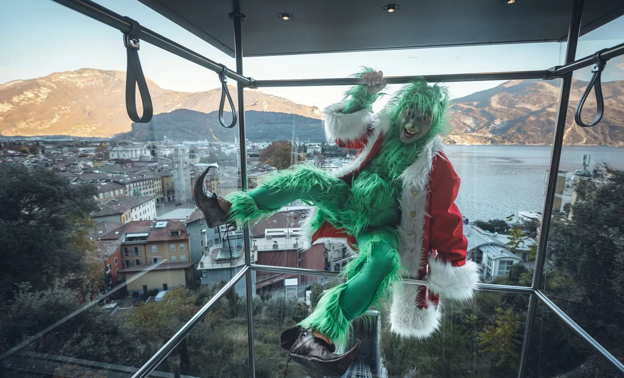 Eine grünliche Figur, die als Weihnachtsmann verkleidet ist, klettert in einen Aufzug mit Blick auf eine Berglandschaft. Die Szene ist lustig und verspielt, mit einer festlichen Atmosphäre.