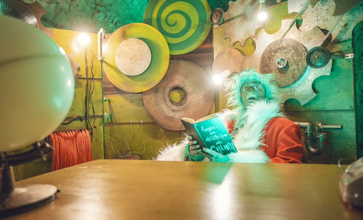 Eine Figur, die als Weihnachtsmann verkleidet ist, liest ein Buch in einer kreativen und bunten Umgebung. Die Wände sind mit Zahnrädern und lebhaften Formen dekoriert.