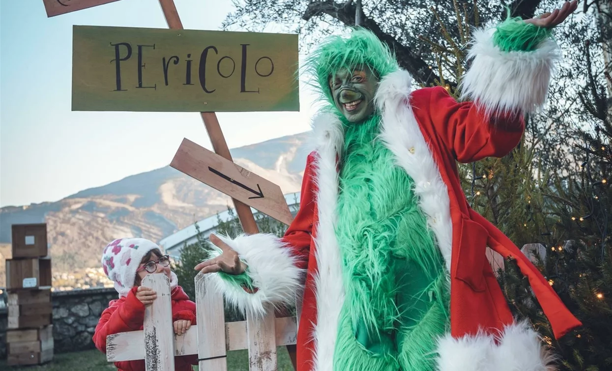 Eine Figur im Grinch-Kostüm lächelt neben einem Mädchen in einer festlichen Umgebung. Es gibt Schilder mit Aufschriften, die dazu einladen, der Grinch zu werden, und warnen vor einer Gefahr.
