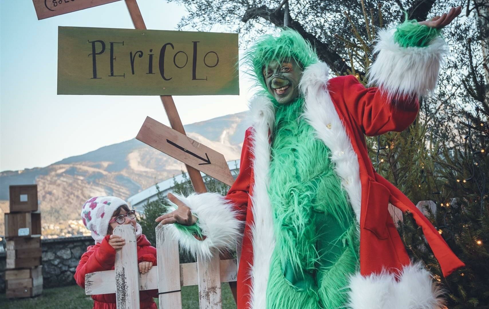 Eine Figur im Grinch-Kostüm lächelt neben einem Mädchen in einer festlichen Umgebung. Es gibt Schilder mit Aufschriften, die dazu einladen, der Grinch zu werden, und warnen vor einer Gefahr.