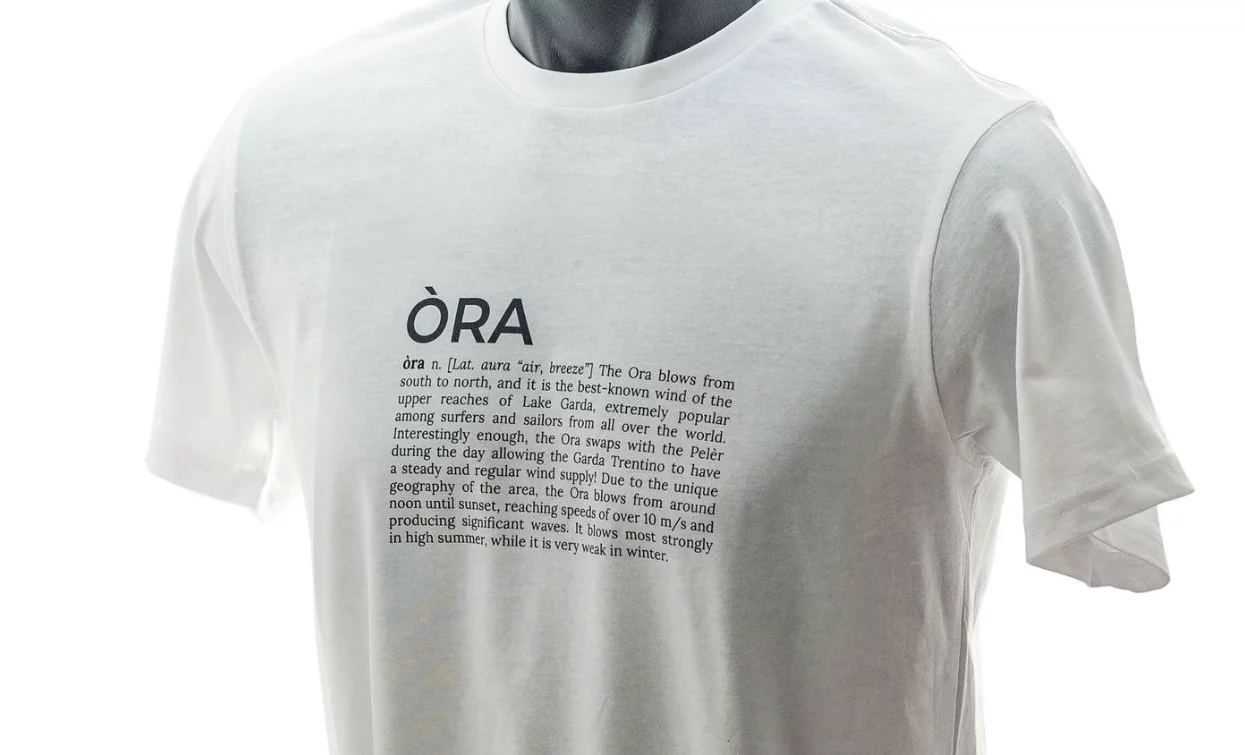 A white t-shirt with the word "ÒRA" printed in large letters. Below, a text that describes the meaning of the word and its connection to the wind and the sea.