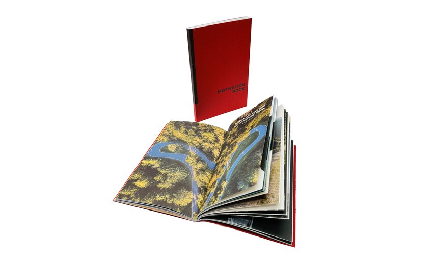 Un libro con copertina rossa è aperto, mostrando pagine illustrate con paesaggi naturali. Le immagini raffigurano un fiume circondato da vegetazione.