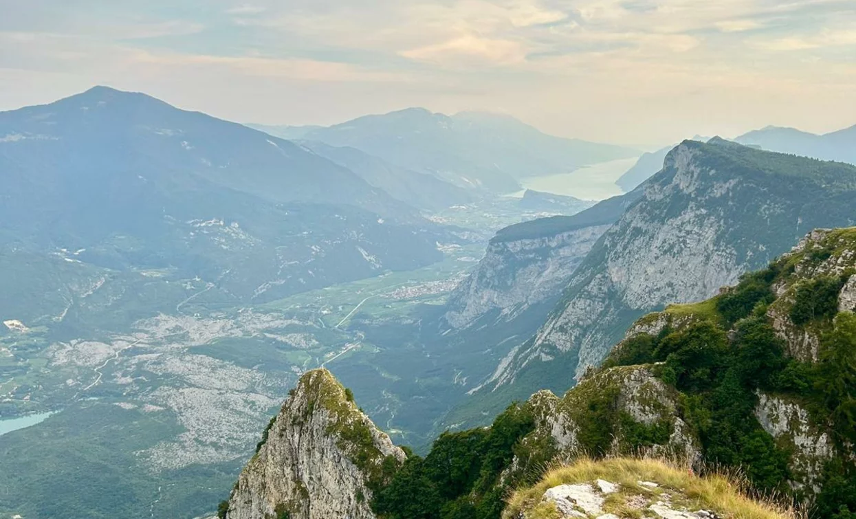 Un paesaggio montano con vette e vallate verdi. Il cielo è nuvoloso e si intravede un lago in lontananza.