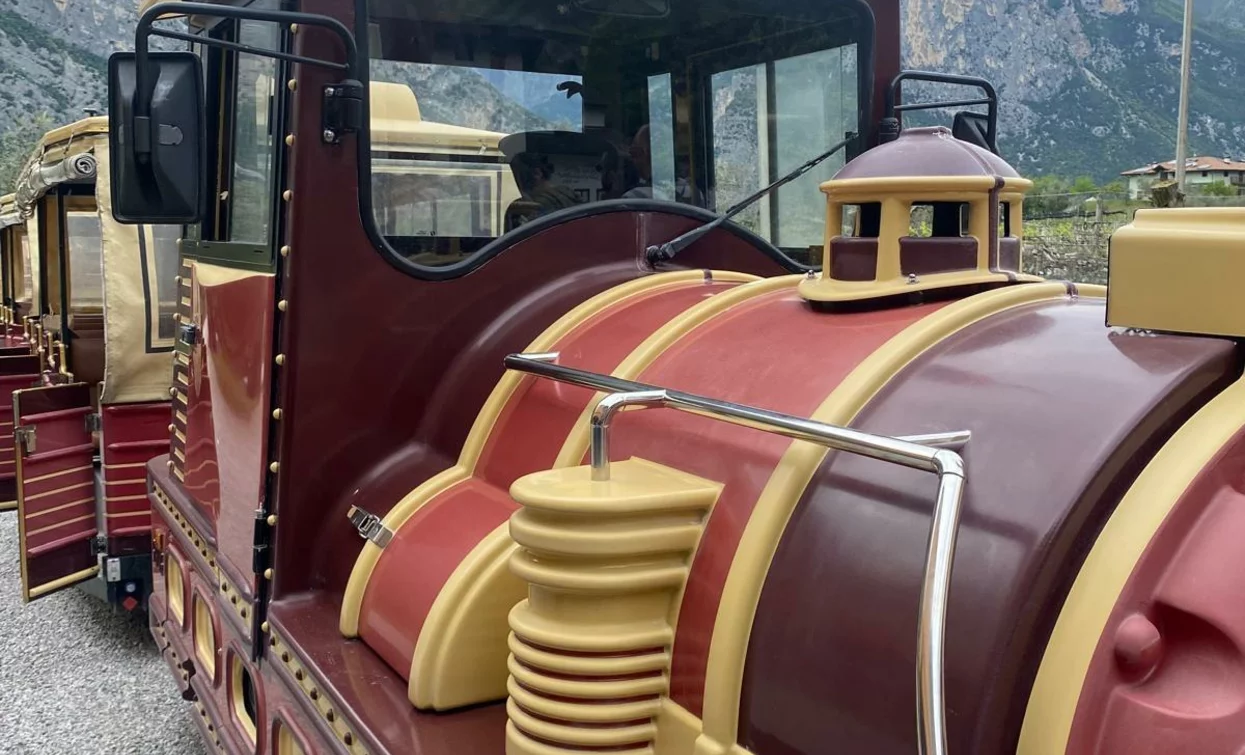Un treno turistico colorato, con dettagli in rosso e giallo. Sullo sfondo si vedono montagne e un cielo nuvoloso.