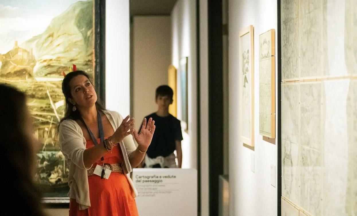Una guida spiega le opere d'arte in un museo. Sullo sfondo, un giovane ascolta attentamente.