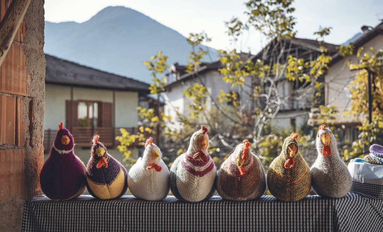 Una fila di galline decorative colorate esposte su un tavolo. Sullo sfondo si vedono case e montagne in un'atmosfera tranquilla.