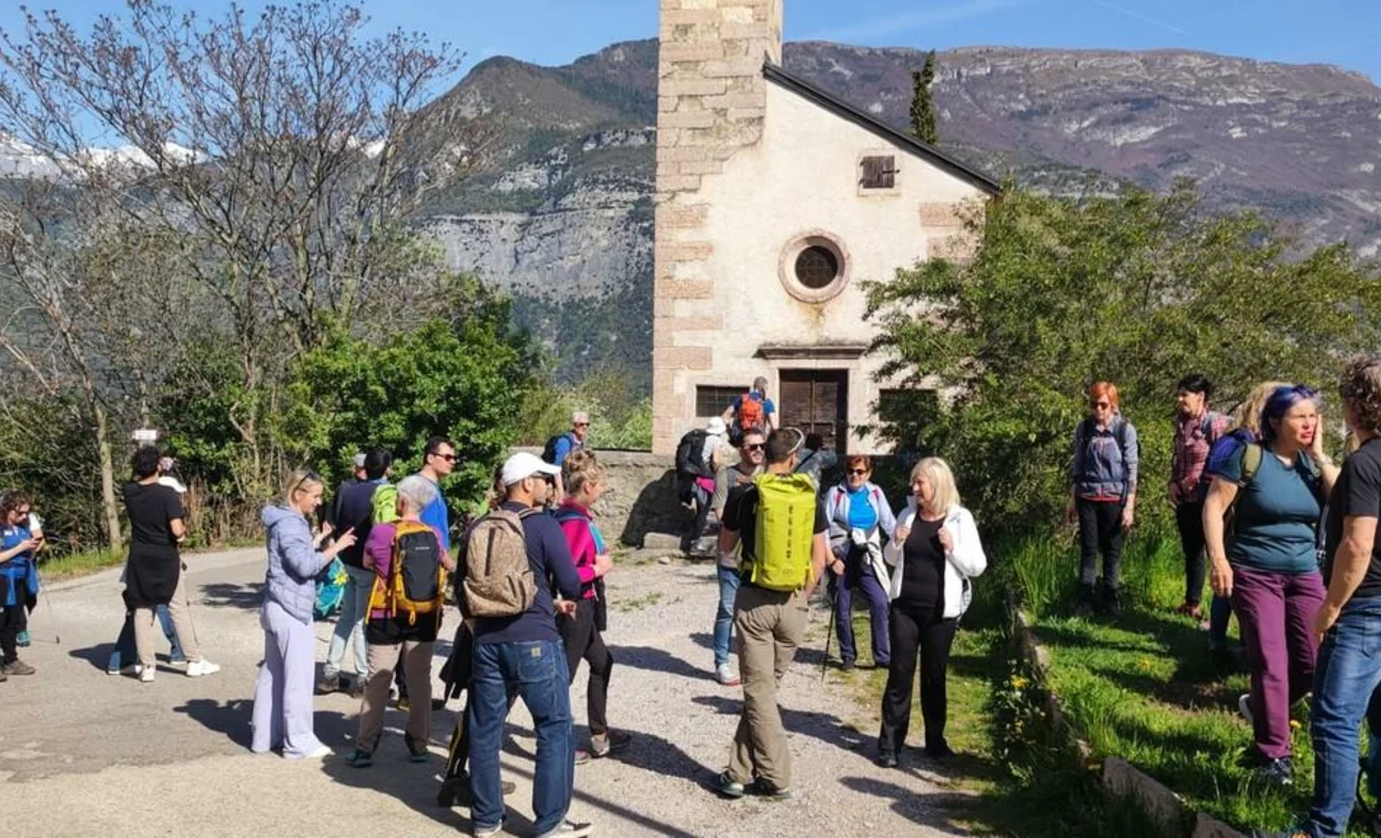 Un gruppo di escursionisti si riunisce davanti a una chiesa in un paesaggio montano. Sullo sfondo si vedono alberi e colline verdi.