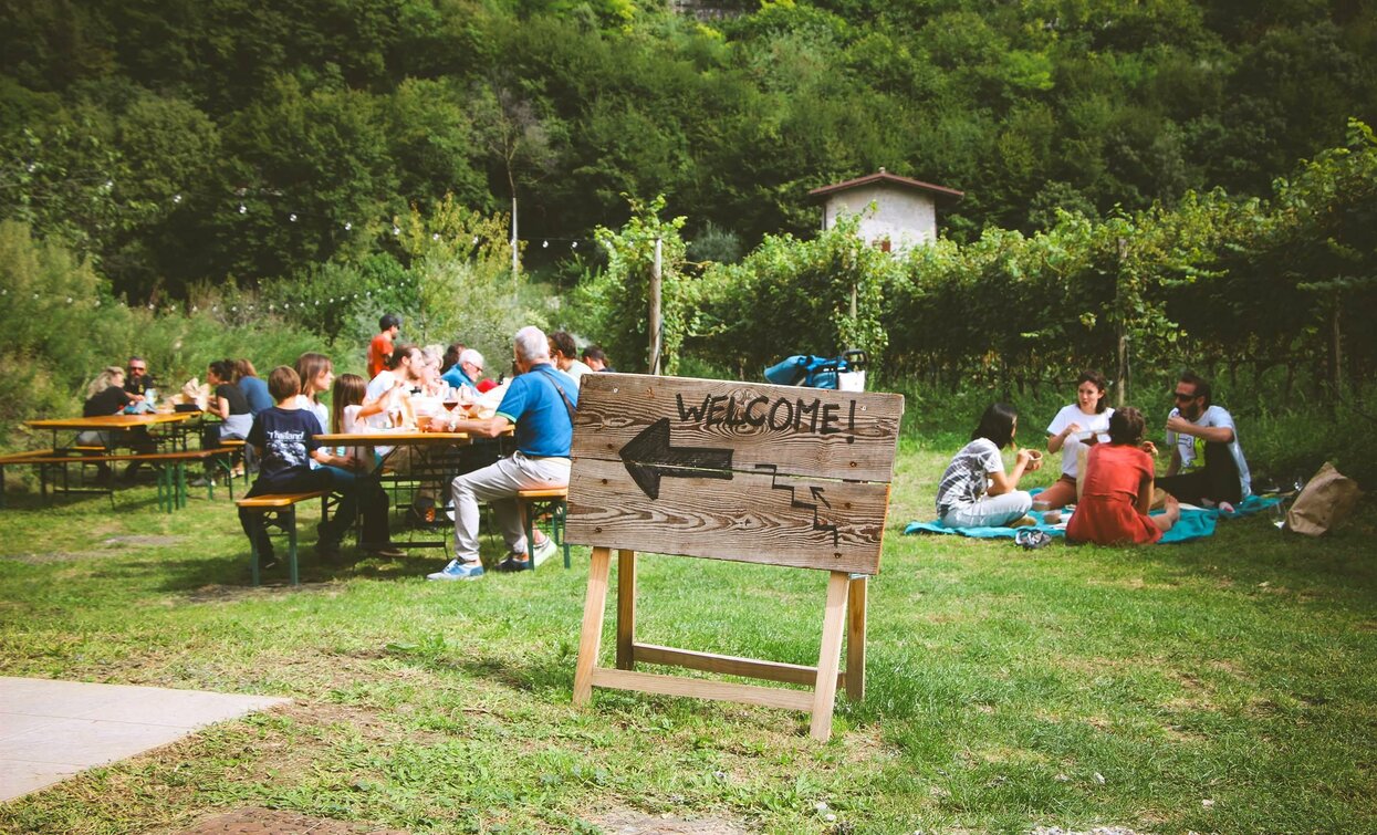 A group of people relaxing in an outdoor garden, sitting at wooden tables. A large wooden sign with the word "WELCOME!" indicates the entrance.