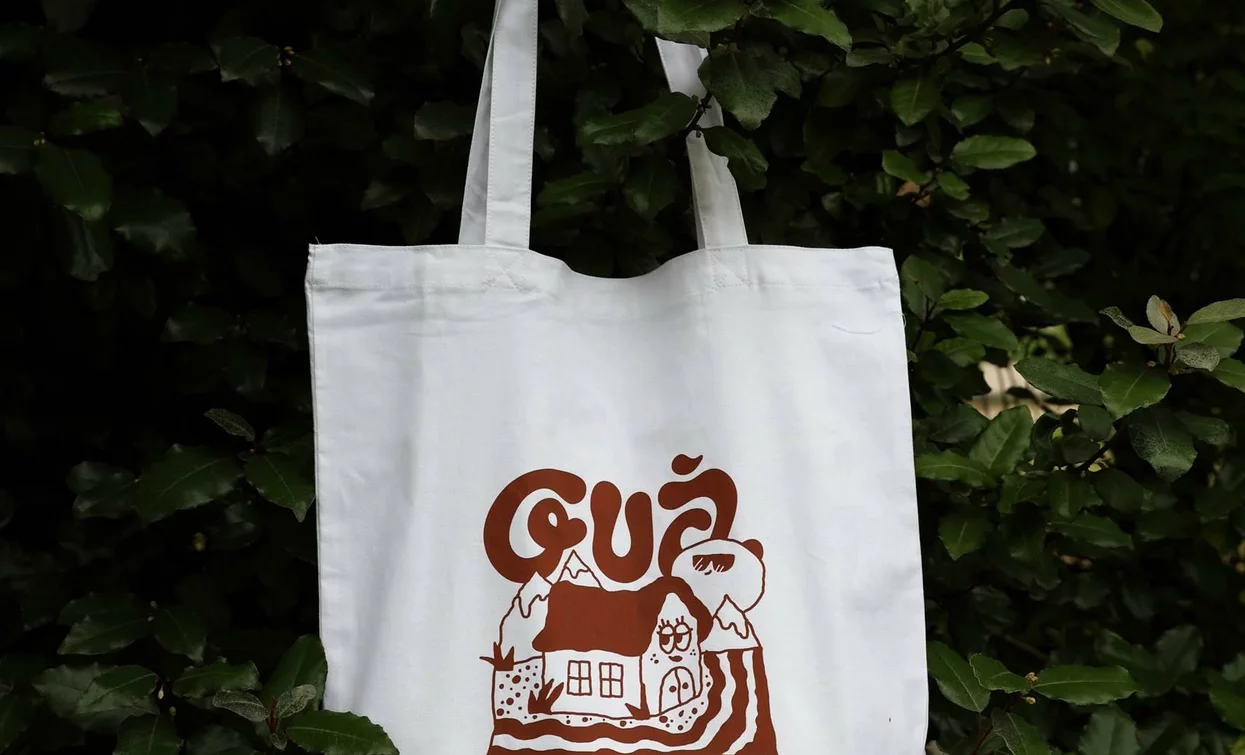 A white canvas bag hanging from a tree, with a simple and attractive design. The writing "Guà" and the image of a farm decorate the bag.