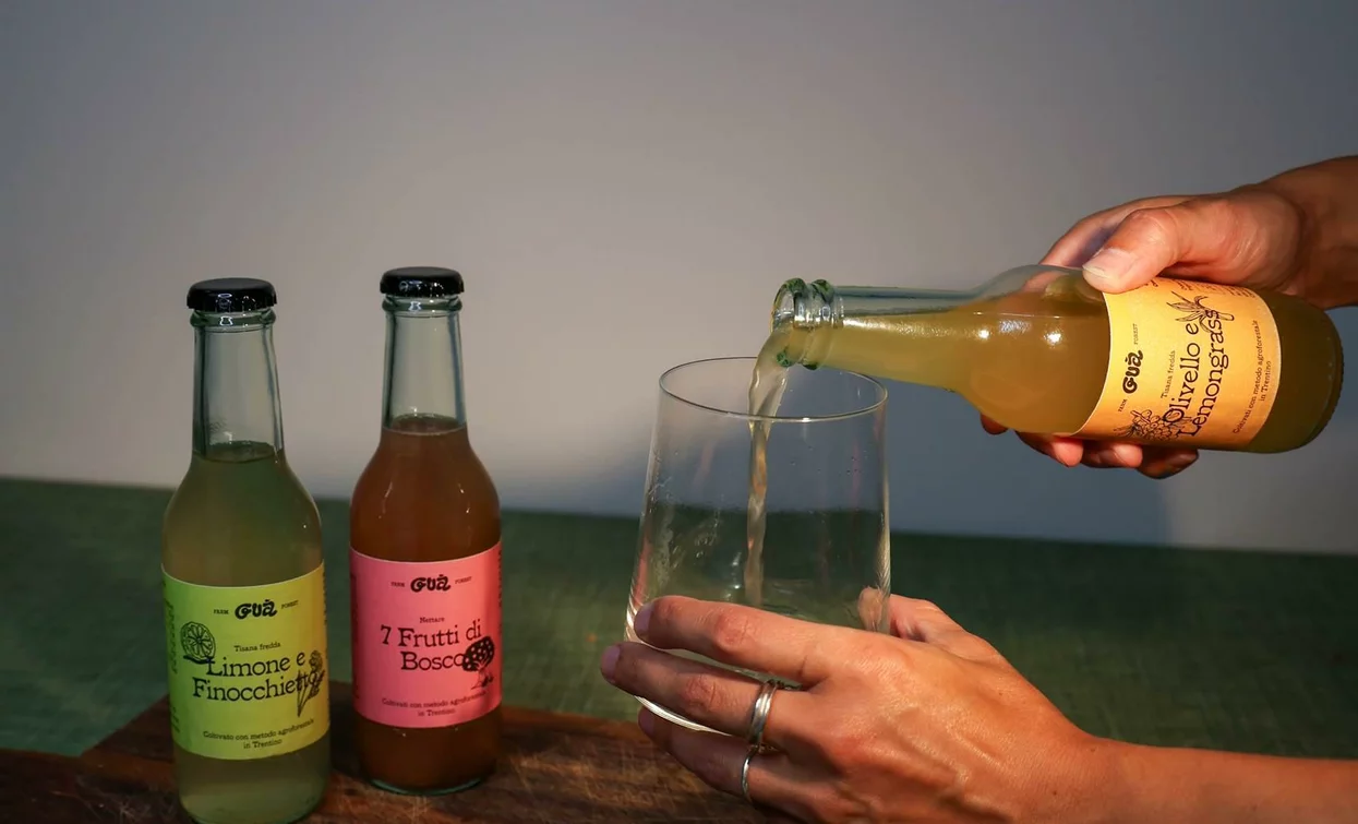 A hand pours a drink into a transparent glass. Two bottles of colorful soda can be seen on a wooden cutting board.