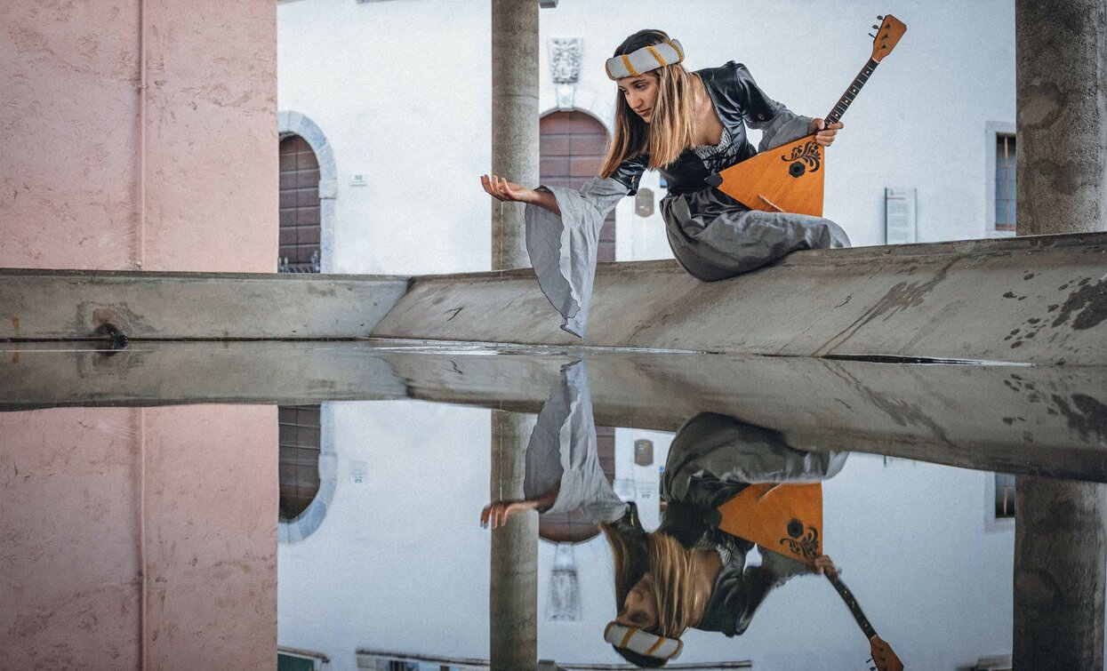Una giovane donna seduta su un muro con un balalaika in mano, riflettendo nell'acqua sottostante. Indossa un abito elegante e sembra immersa nei suoi pensieri.