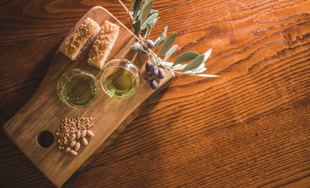 Ein Holzbrett präsentiert zwei Gläser Olivenöl, knusprige Snacks und einen Zweig Olivenbaum. Die Oberfläche ist mit elegantem Holz überzogen, das die Farben der Zutaten betont.